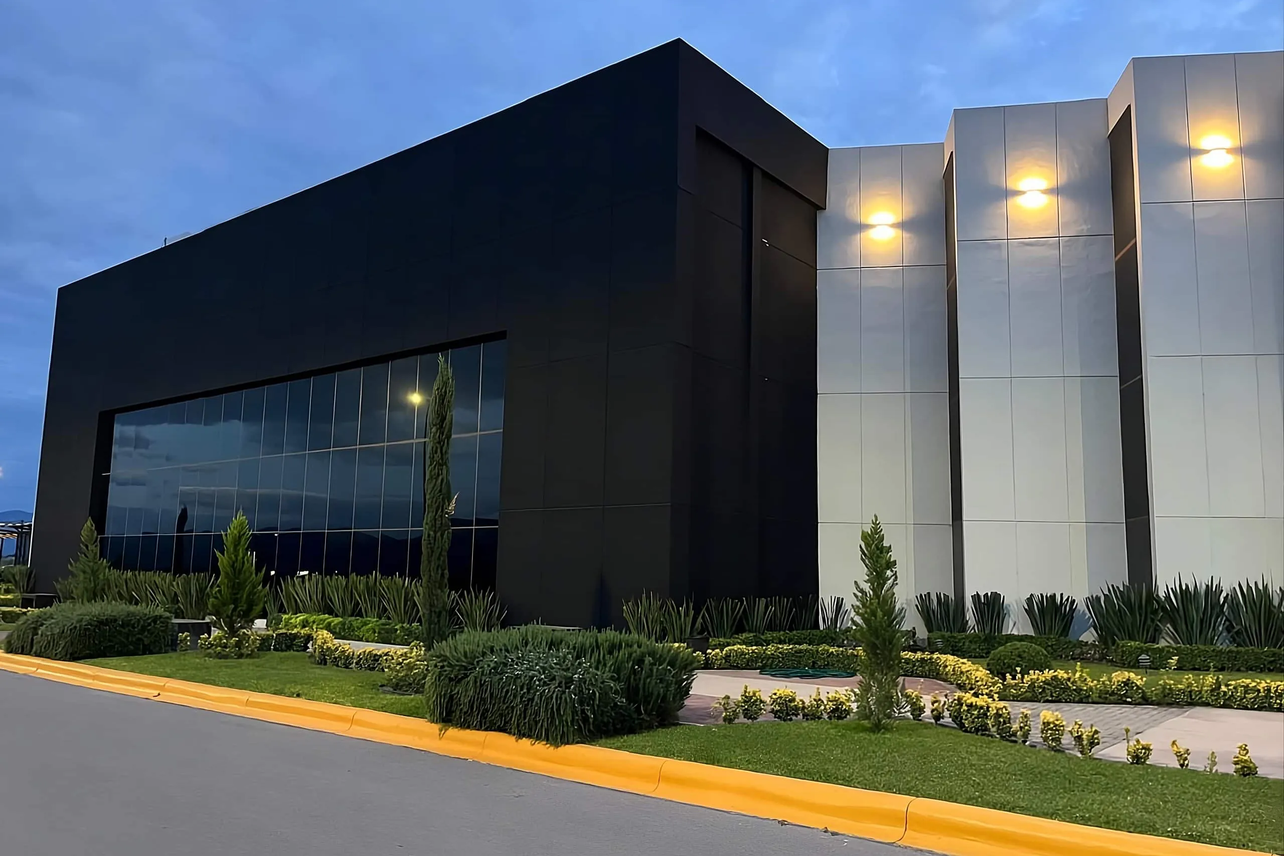 Black and white facade of the Grupo Arga plant at dusk, with its wall lights on